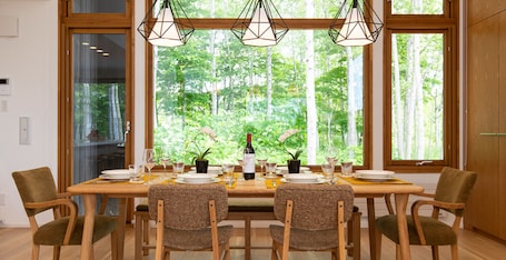 A dining area with a table set for six, surrounded by chairs, with a view of trees through large windows.