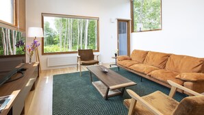 A modern living room with a brown sofa, a wooden coffee table, a chair, and a TV on a wooden stand. Large windows offer views of trees. - Luxury 5 Star 3 Bedroom Chalet, Niseko Chalet 1005 (Niseko)