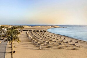 Una playa con hileras de sombrillas y palmeras, una orilla arenosa y un mar tranquilo.