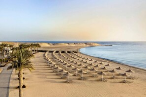A beach with rows of umbrellas and palm trees, a clear sky, and a calm sea. - JAZ Costa Mares - Adults Only 16+ (Marsa Alam)