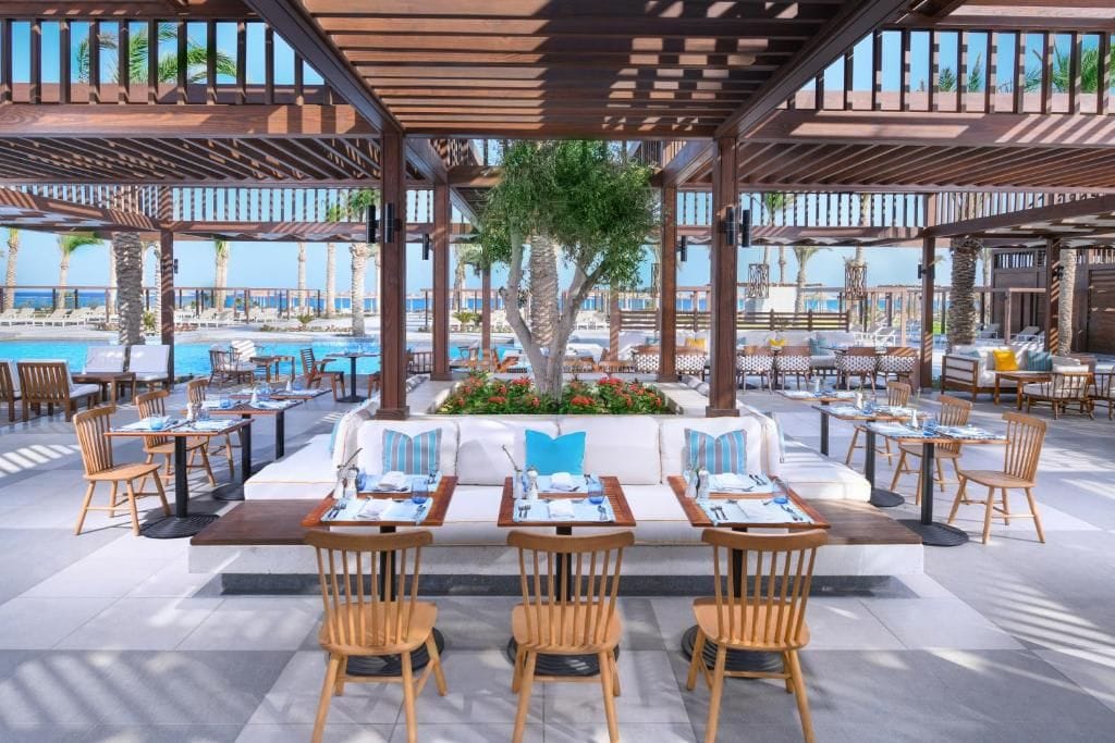 An outdoor dining area with wooden tables and chairs, a central seating area with cushions, and a view of the pool and palm trees.