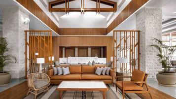 Un intérieur moderne avec un plafond haut, un espace salon central avec un canapé et des fauteuils, et une cuisine en arrière-plan.