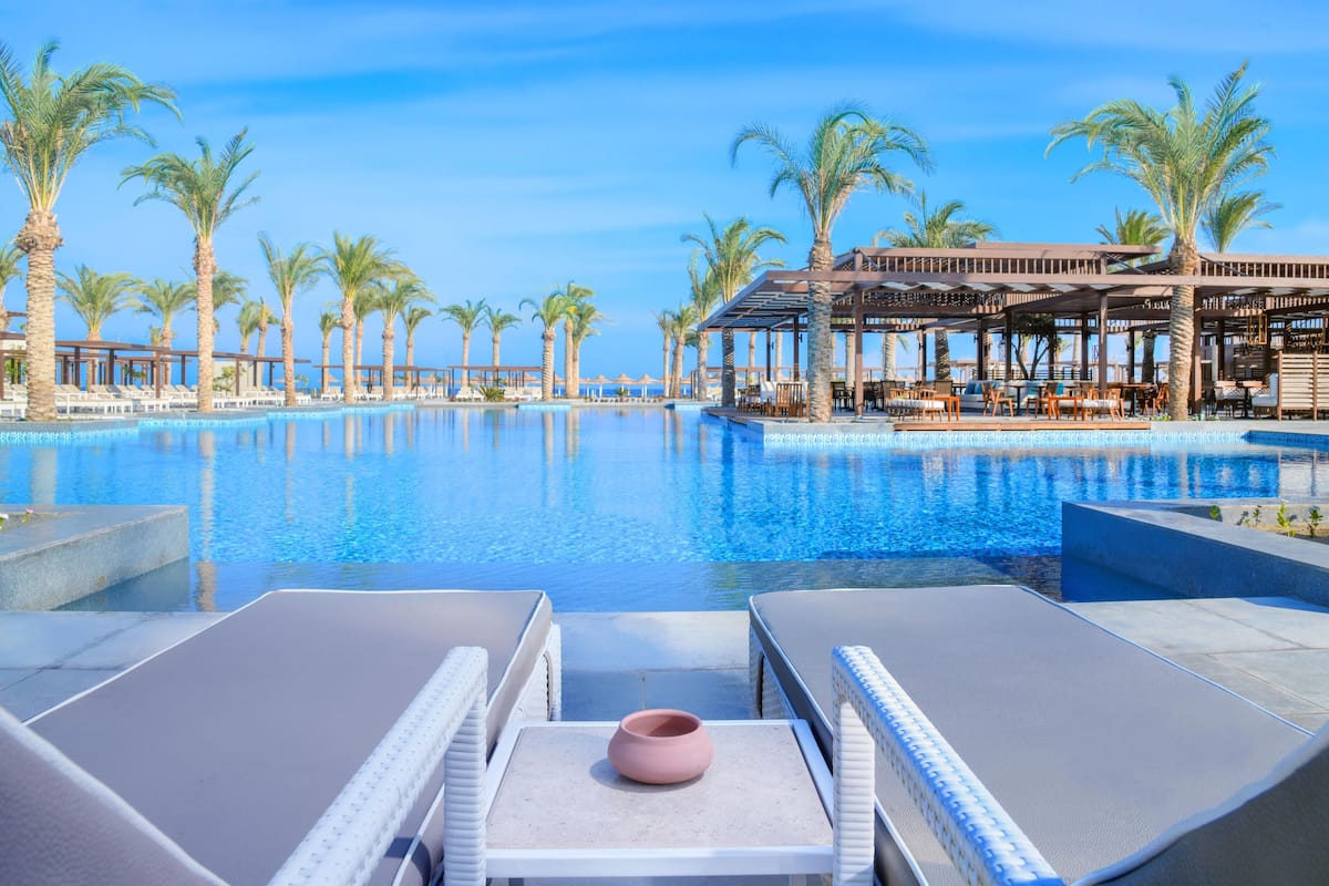 Ein Poolbereich mit Liegestühlen und einem Tisch, umgeben von Palmen und blauem Himmel.