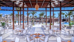 Un área de comedor al aire libre con mesas y sillas, una piscina al fondo y palmeras.