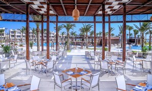An outdoor dining area with tables and chairs, a pool in the background, and palm trees. - JAZ Costa Mares - Adults Only 16+ (Marsa Alam)