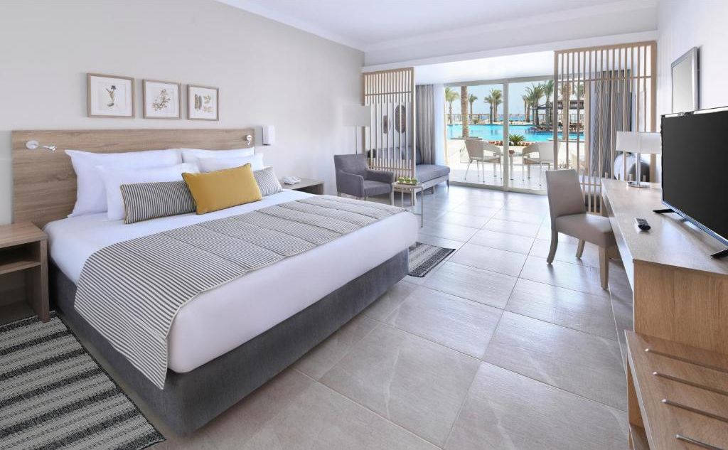 A hotel room with a bed, a desk, a chair, a TV, and a view of a pool.