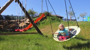 Lapsi keinussa, taustalla ruohikkoalue, liukumäki ja puinen rakennelma.