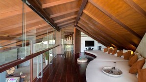 A modern interior with a high ceiling, a curved seating area with cushions, and a glass railing overlooking a deck.