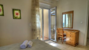 Une chambre à coucher comprenant un lit, une commode en bois, une chaise et une porte-fenêtre coulissante donnant sur un balcon.