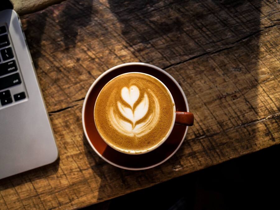 A cup of coffee with a heart design on a wooden table next to a laptop.
