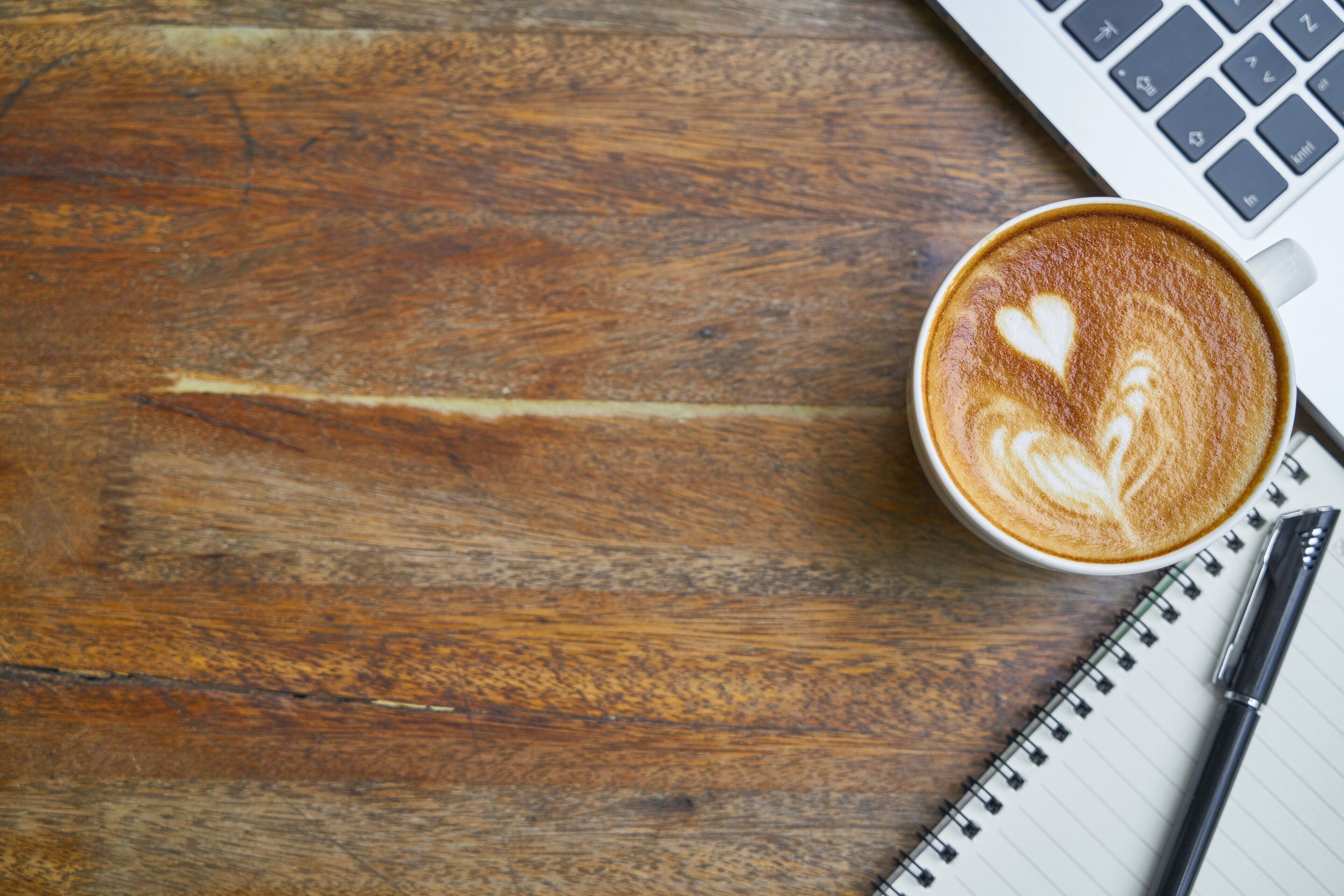 A wooden table with a cup of coffee featuring latte art, a notebook, and a pen.
