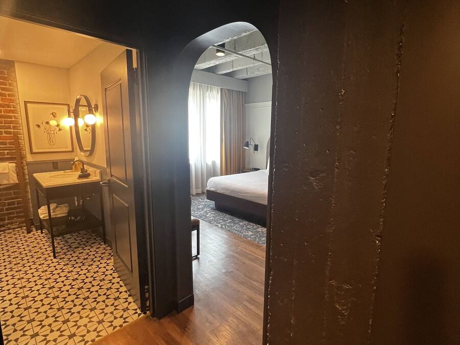 A hotel room with a wooden floor, a bed with a white mattress, a small table with a chair, a mirror, and a picture on the wall.
