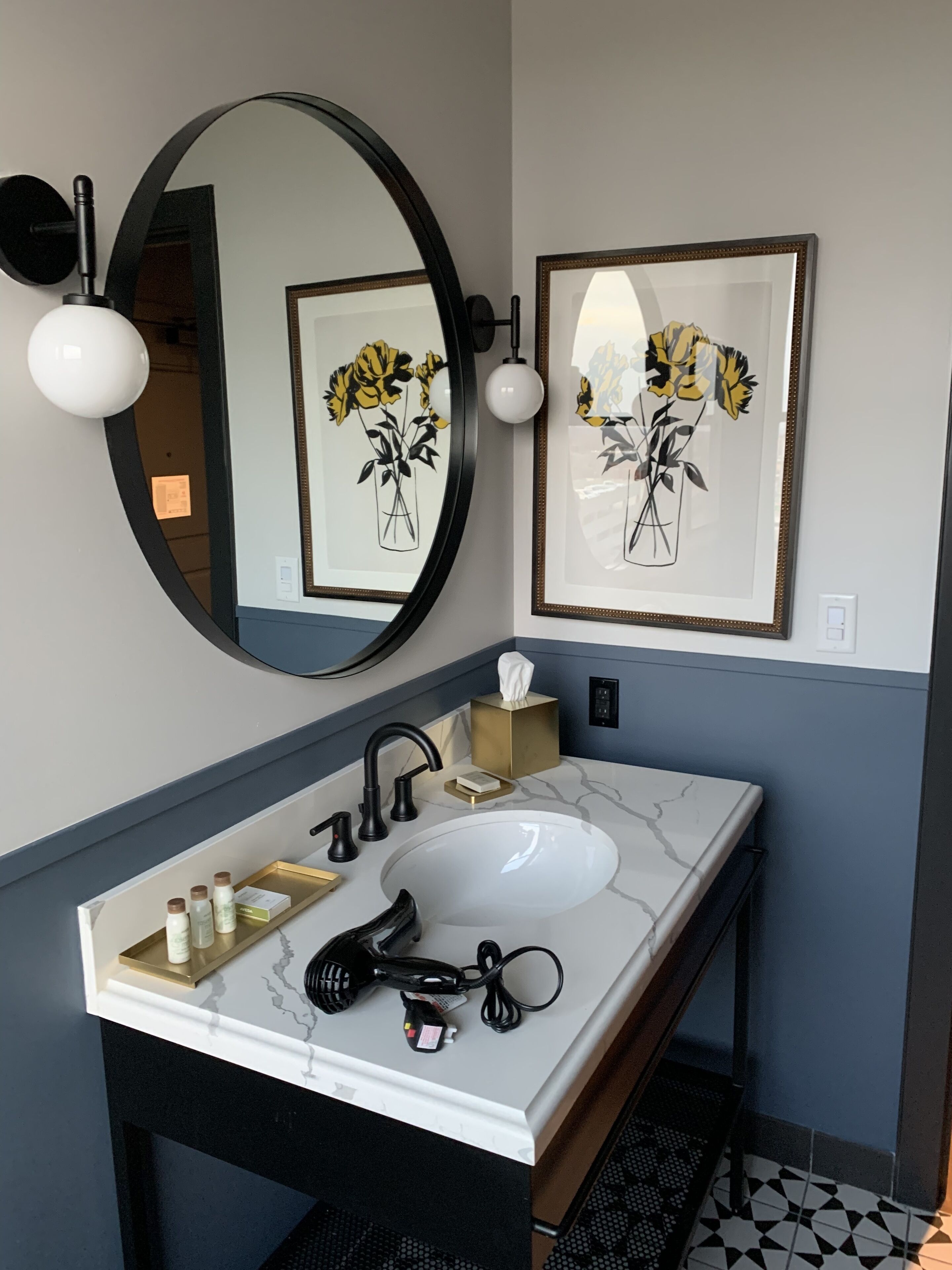 Une salle de bain avec un lavabo en marbre blanc, un robinet noir et un miroir avec un cadre doré. Des tableaux encadrés représentant des fleurs sont accrochés au mur, et une boîte de mouchoirs est posée sur le comptoir.