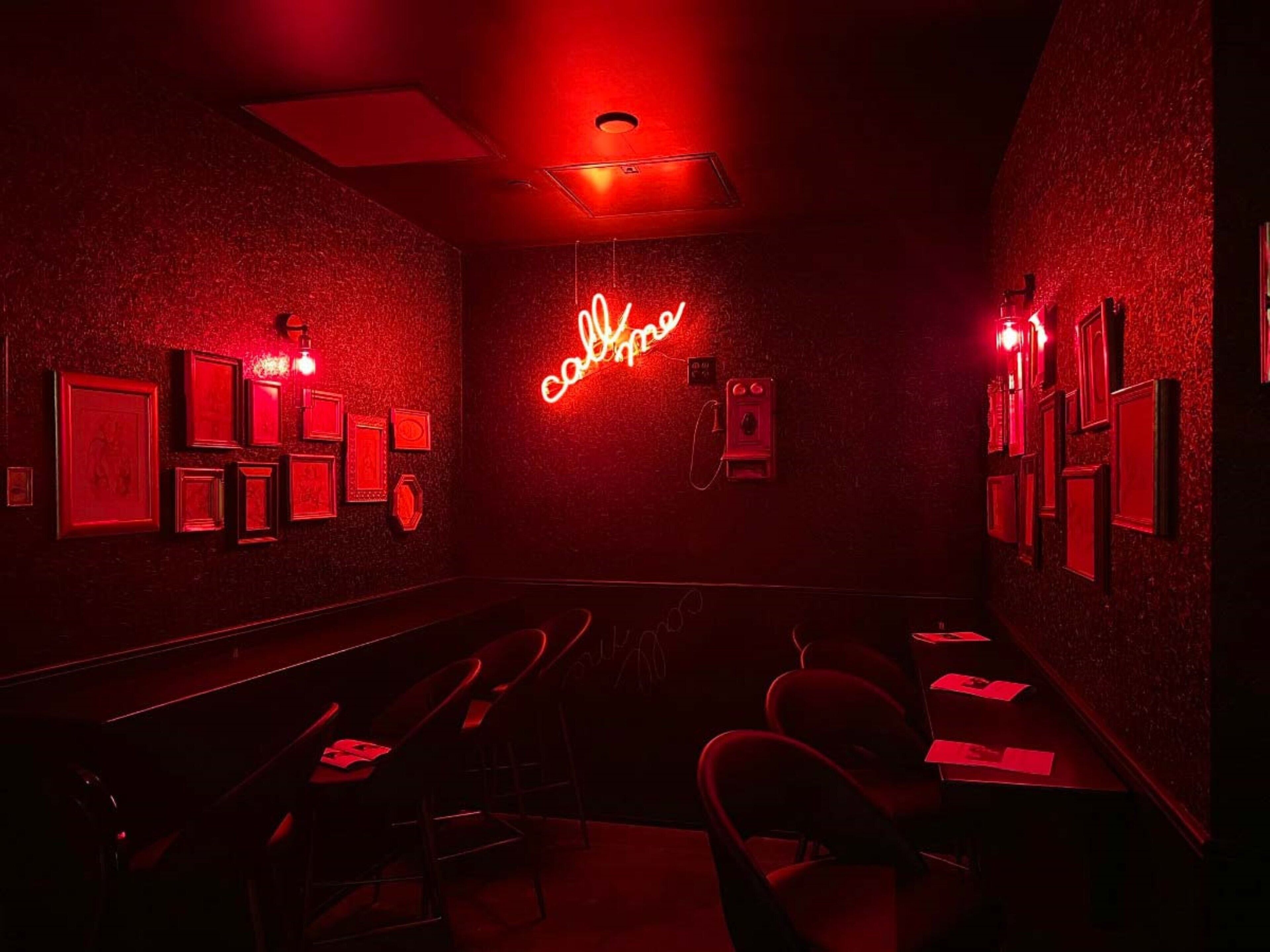 A dimly lit room with red ambient lighting, a neon sign reading "call me", and several framed pictures on the walls.