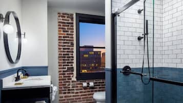 Un baño con pared de ladrillo, ventana con vista a la ciudad, lavabo blanco y zona de ducha con mampara de vidrio.