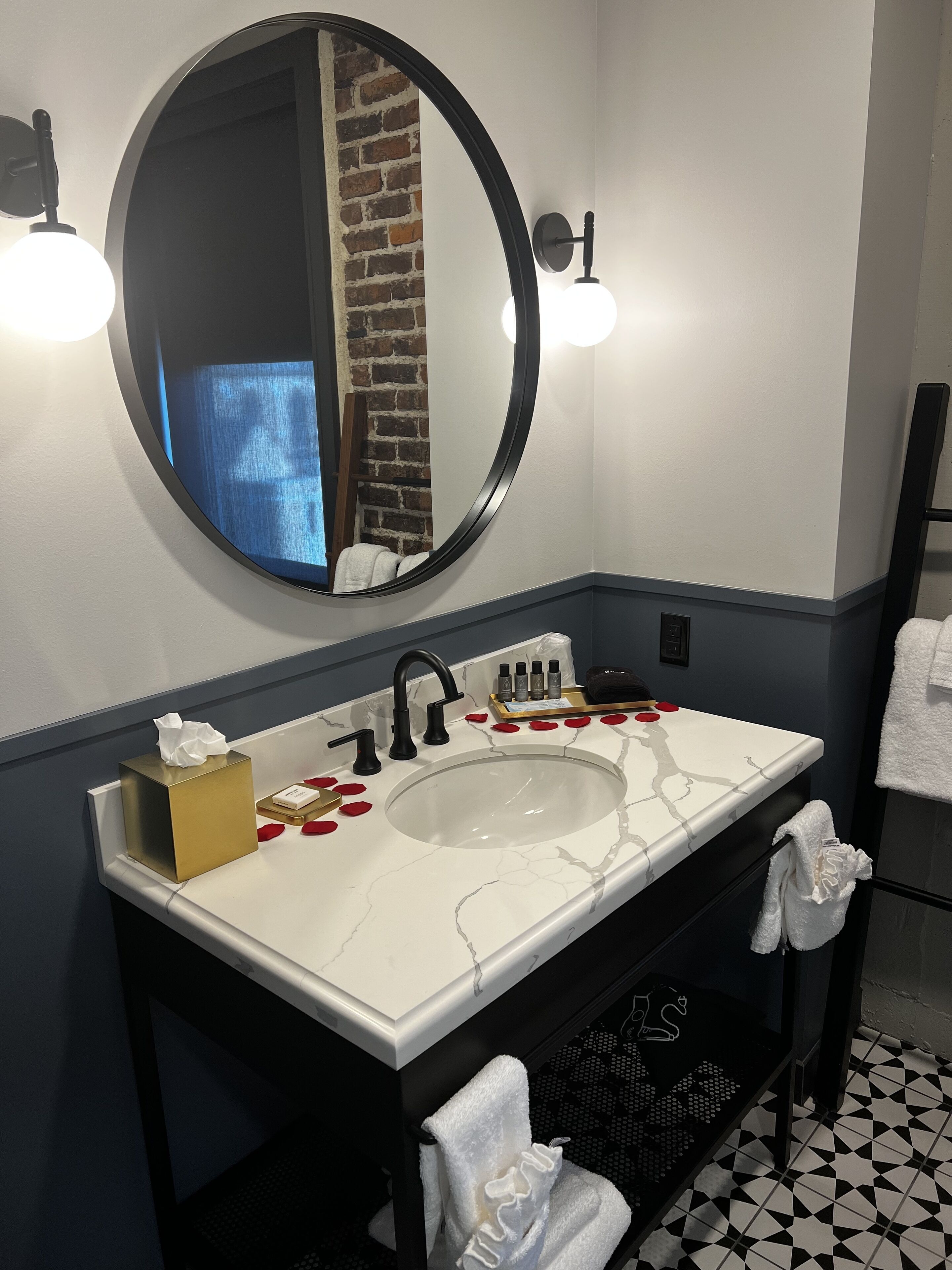 A bathroom with a white marble sink, black faucet, and a round mirror. There are rose petals on the counter, a tissue box, and a towel rack with towels.