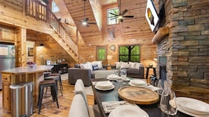A spacious cabin interior with a wooden staircase, a dining area with a round wooden table, and a stone fireplace.