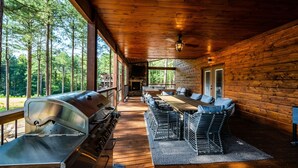 Een overdekt terras met een barbecue, houten tafels en stoelen, omgeven door een bos.