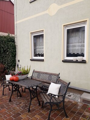 A patio with a table and two benches, a potted plant, and a wall with two windows. - Ferienwohnung Erika, Deutschland (Blankenburg (Harz))