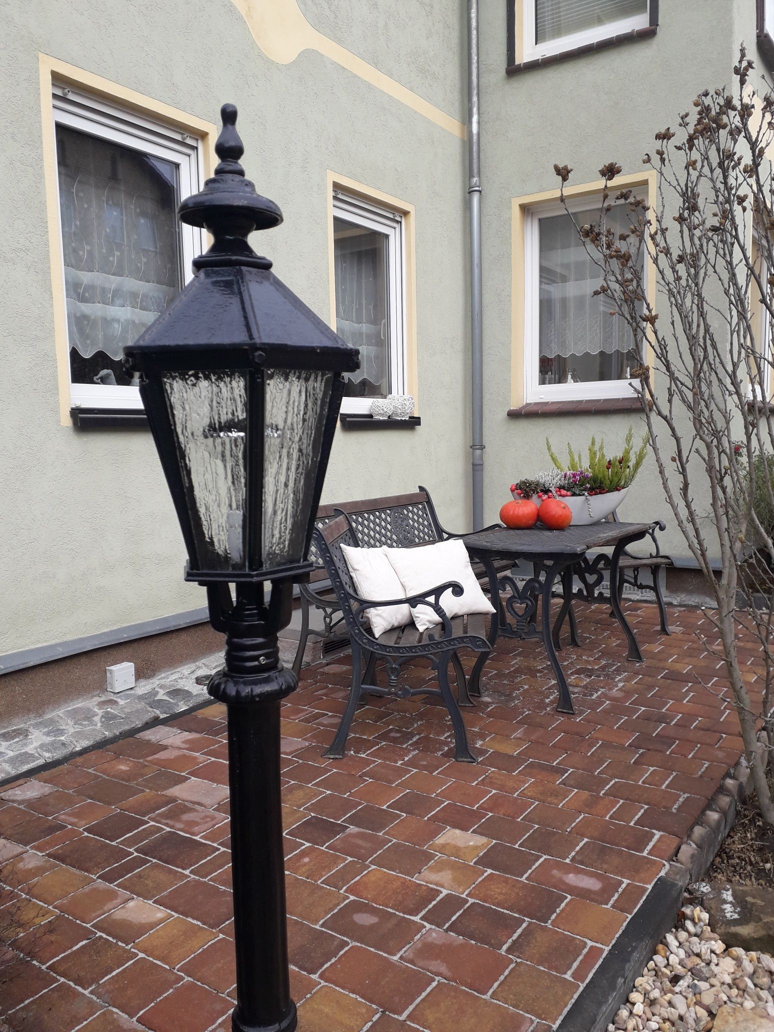 Een terras van stenen met een zwarte lantaarnpaal, een bank en een tafel met een potplant.