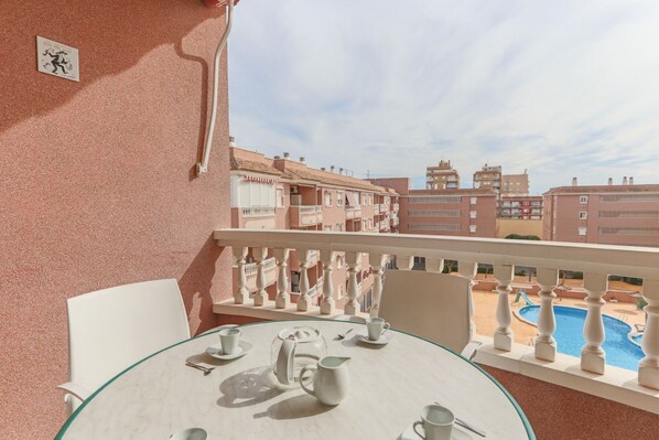 A balcony with a glass table set for tea, overlooking a swimming pool and residential buildings.