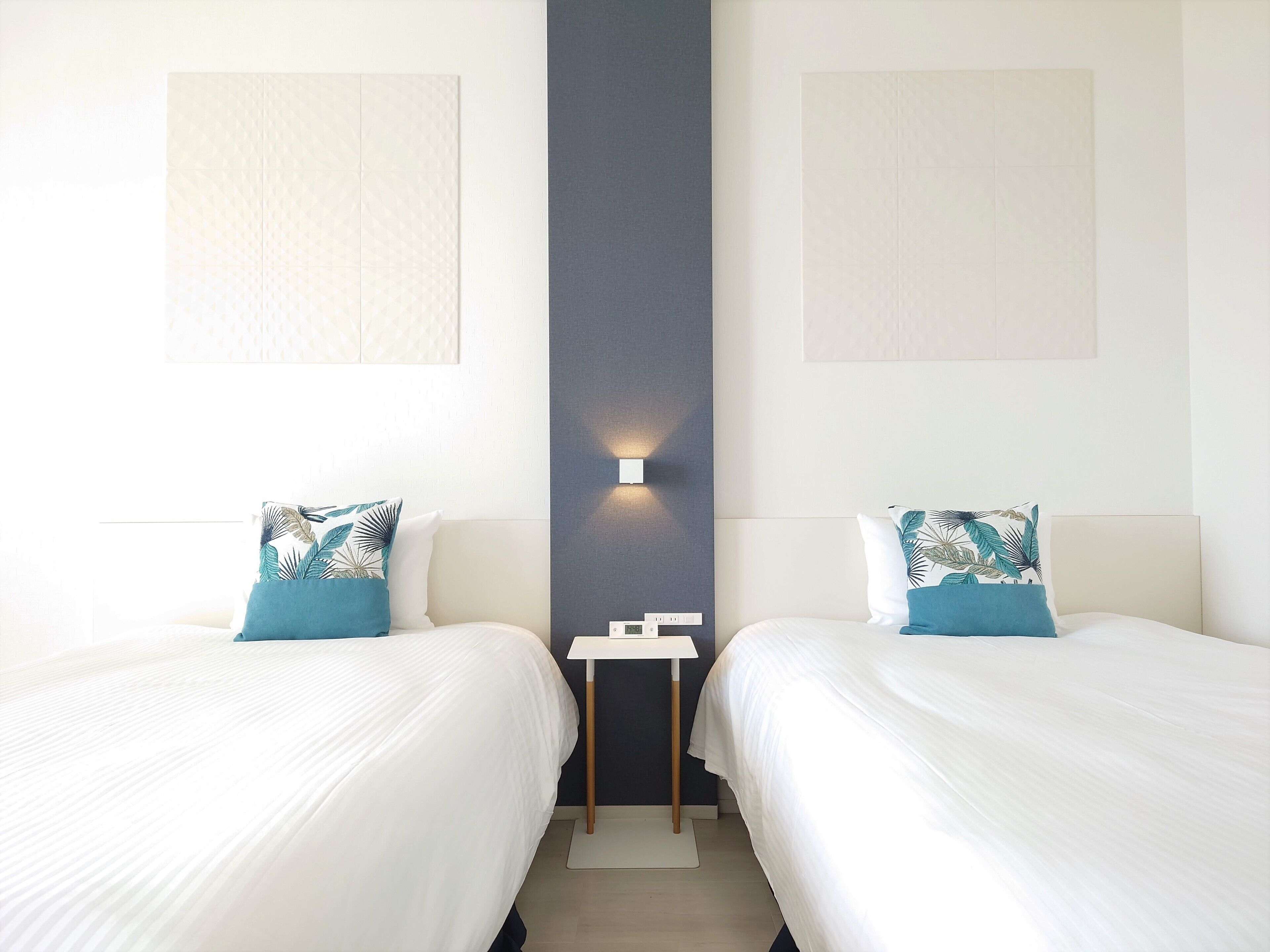 A hotel room with two beds, each with a white bedspread and a blue pillow with a leaf pattern. There is a small table between the beds with a white lamp on it.