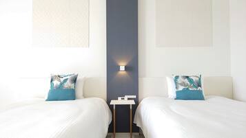 A hotel room with two beds, each with a white bedspread and a blue pillow with a leaf pattern. There is a small table between the beds with a white lamp on it.