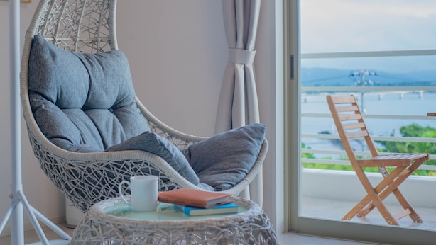 Une chaise en osier avec des coussins, une petite table avec un livre et une tasse, et une vue sur un pont et la verdure, le tout visible à travers une fenêtre.