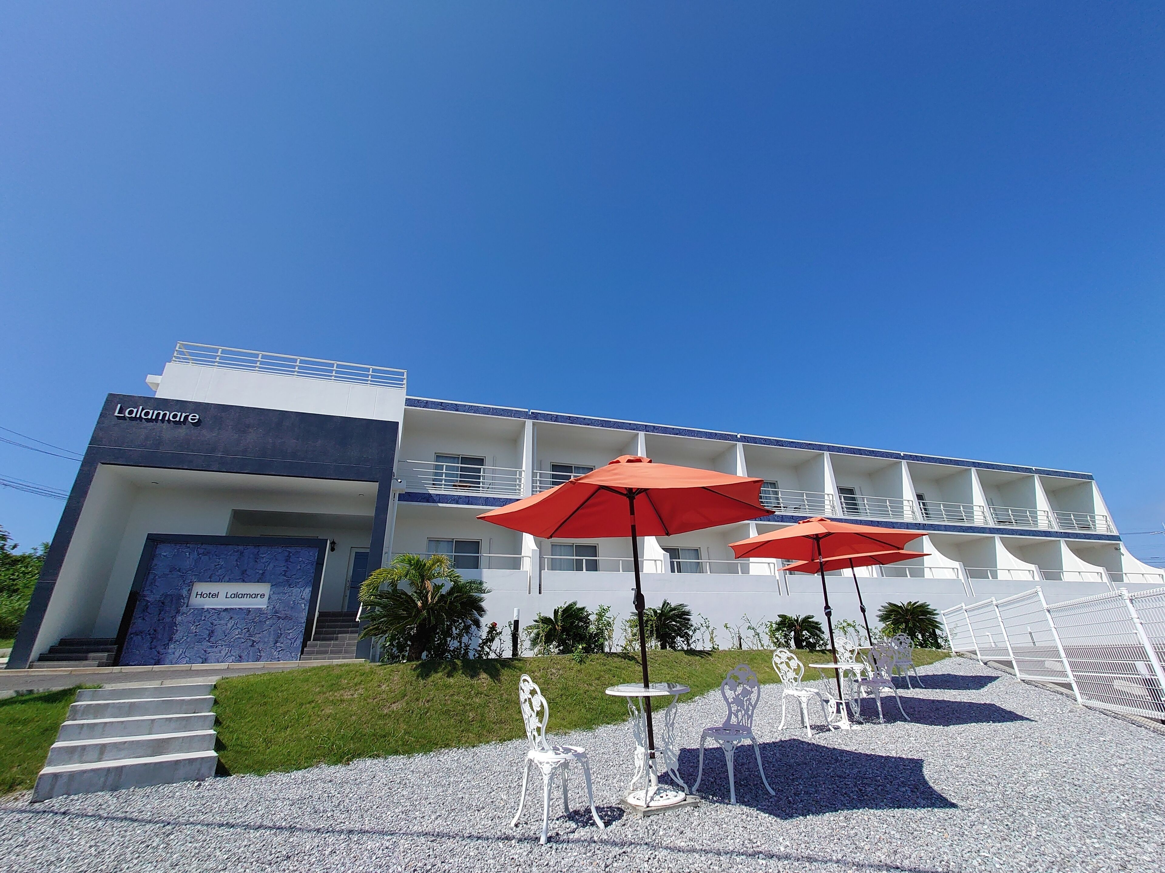 A hotel named 'Lalamare' with a modern design, featuring a blue and white facade, outdoor seating with red umbrellas, and a gravel patio area.