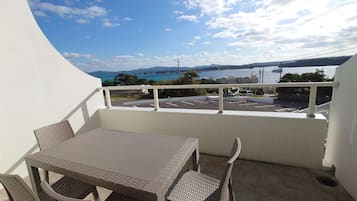 A balcony with a table and chairs, overlooking a parking area and a body of water with mountains in the distance.