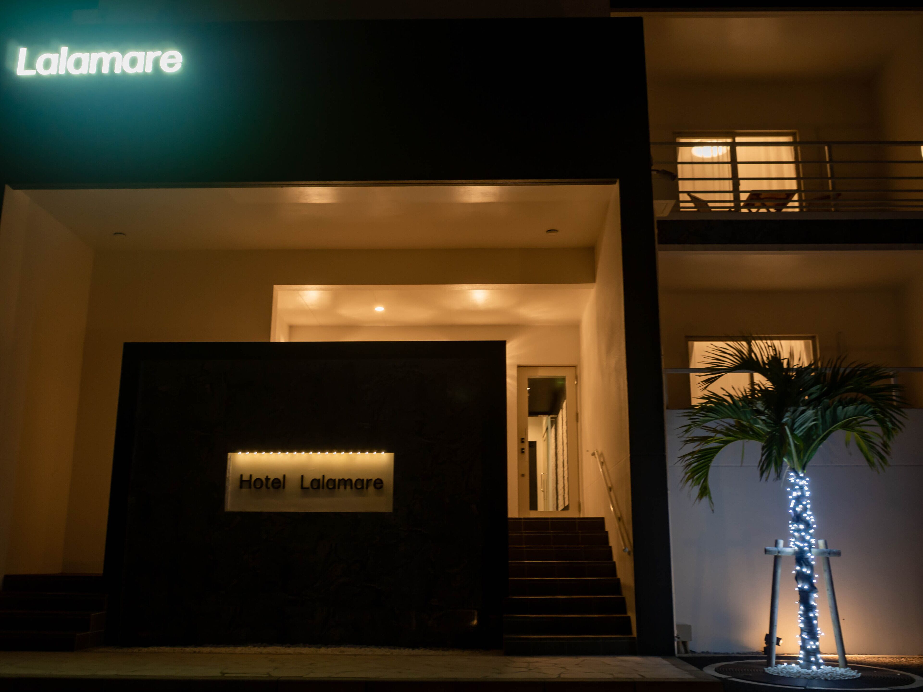 The exterior of Hotel Lalamare at night with a signboard, a staircase, and a palm tree with lights.
