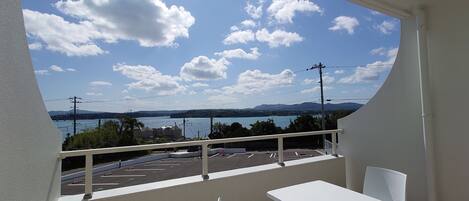 A balcony with white chairs and a table, overlooking a parking area and a body of water with mountains in the distance.