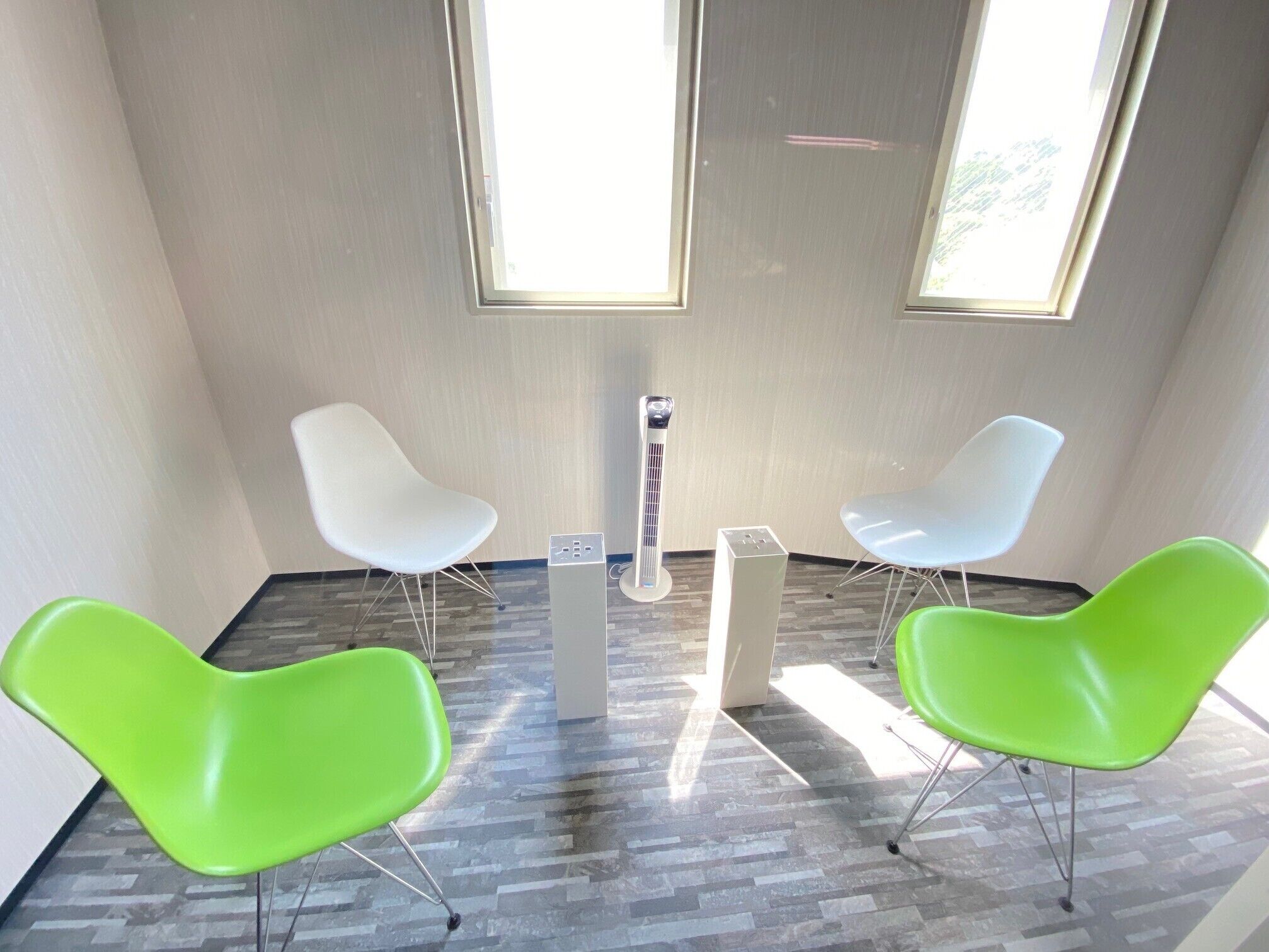 A modern room with two white chairs and two green chairs, a central air conditioning unit, and large windows allowing natural light.