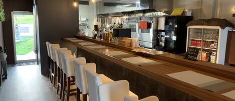 A modern bar area with a long counter, bar stools, and a view of the kitchen in the background.