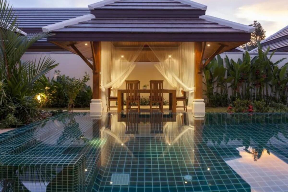 A modern resort with a swimming pool, a covered seating area with chairs, and lush greenery.