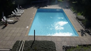 Une piscine rectangulaire aux eaux d’un bleu limpide, entourée de chaises longues et de verdure, avec une chaîne de montagnes en arrière-plan.