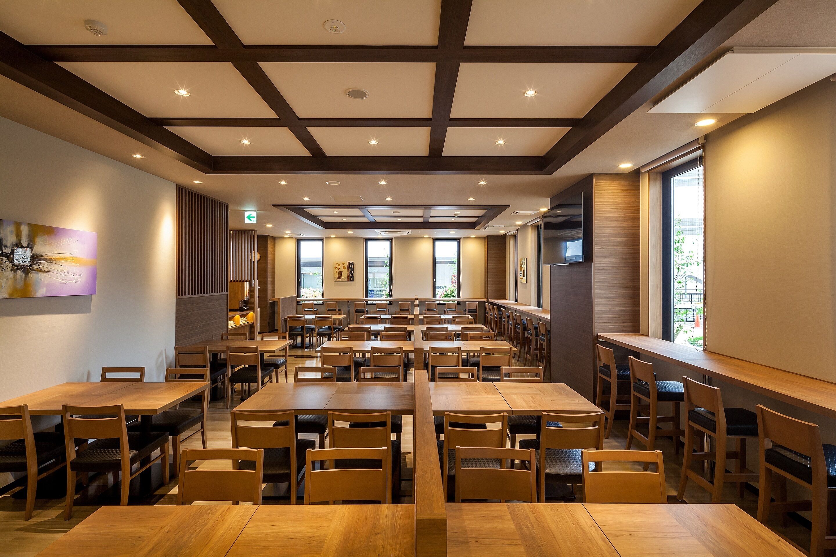 A spacious dining area with multiple wooden tables and chairs, large windows, and a modern ceiling design.