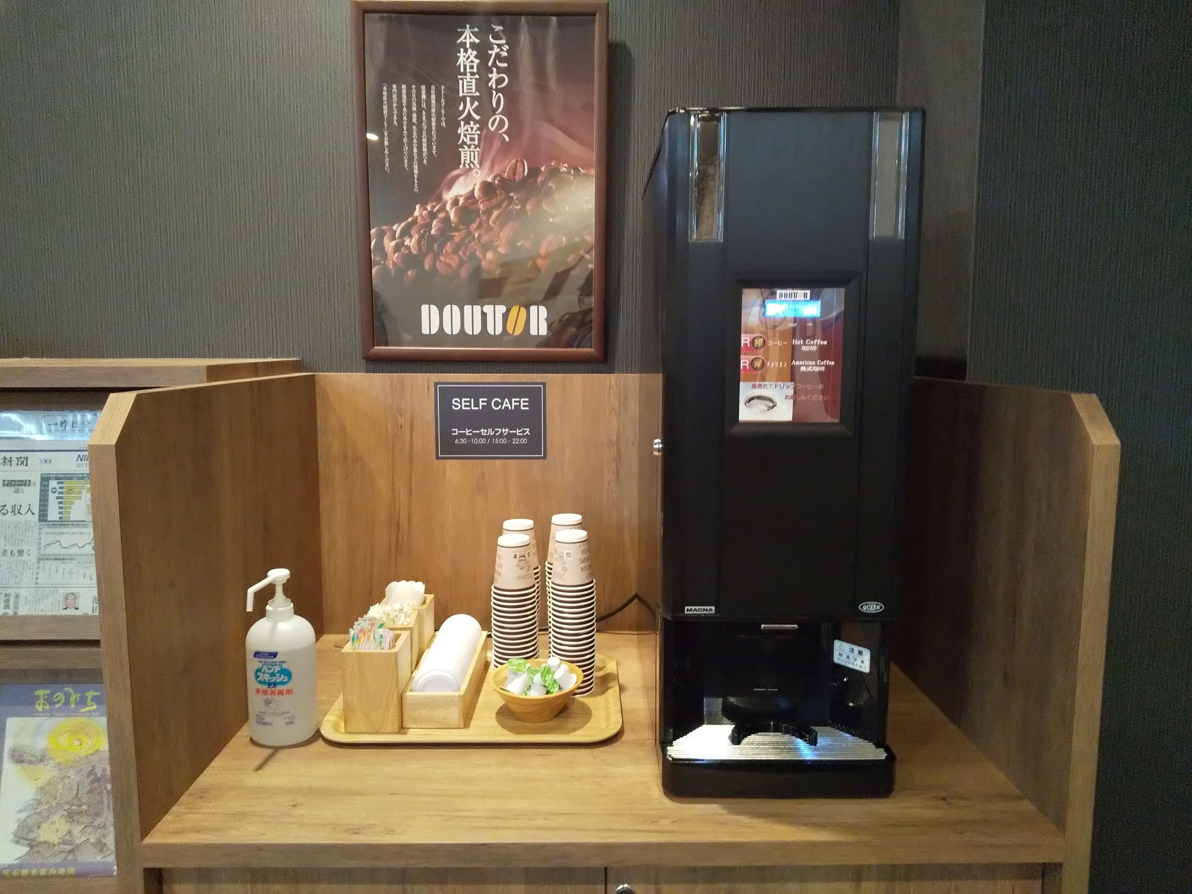 A self-service coffee station with a coffee machine, disposable cups, and a tray with coffee-related items.