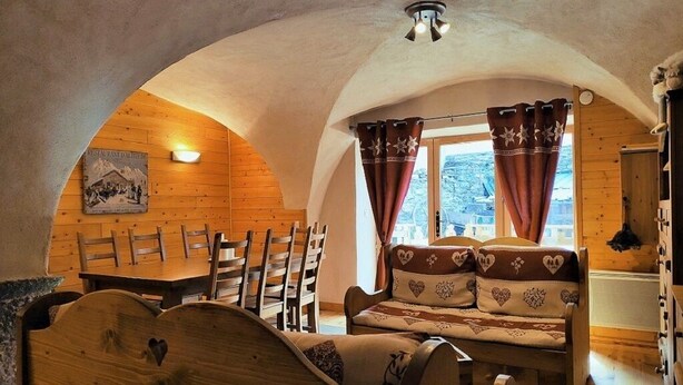 Ein gemütlicher Raum mit Holzwänden und schrägen Decken, ausgestattet mit einem hölzernen Bett, einer Couch mit Kissen mit Herzmuster, einem Esstisch mit Stühlen und einem Fenster mit Vorhängen.
