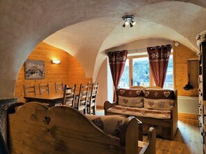 Une pièce chaleureuse aux murs en bois et au plafond en pente, comprenant un lit en bois, un canapé agrémenté de coussins à motifs en forme de cœur, une table à manger avec des chaises et une fenêtre avec des rideaux.