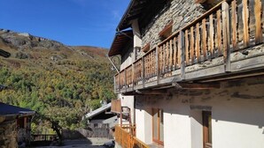 Ein traditionelles Steinhaus mit Holzbalkon, Kiesweg und Bergelandschaft im Hintergrund.