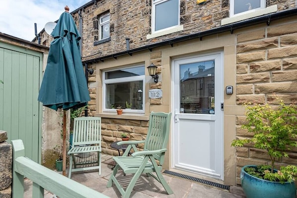 Une maison en pierre avec une porte verte, deux fenêtres et une petite terrasse avec une table et des chaises.