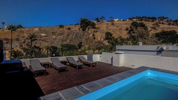 A rooftop pool with lounge chairs, a wooden deck, and a view of a hillside with trees and buildings. - Luxury 2 Bedroom With Roof Deck and Pool Dewaterkant (Cape Town)