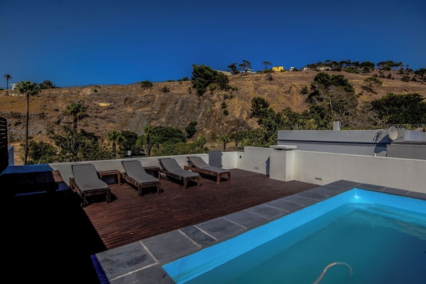 A rooftop pool with lounge chairs, a wooden deck, and a view of a hillside with trees and buildings.