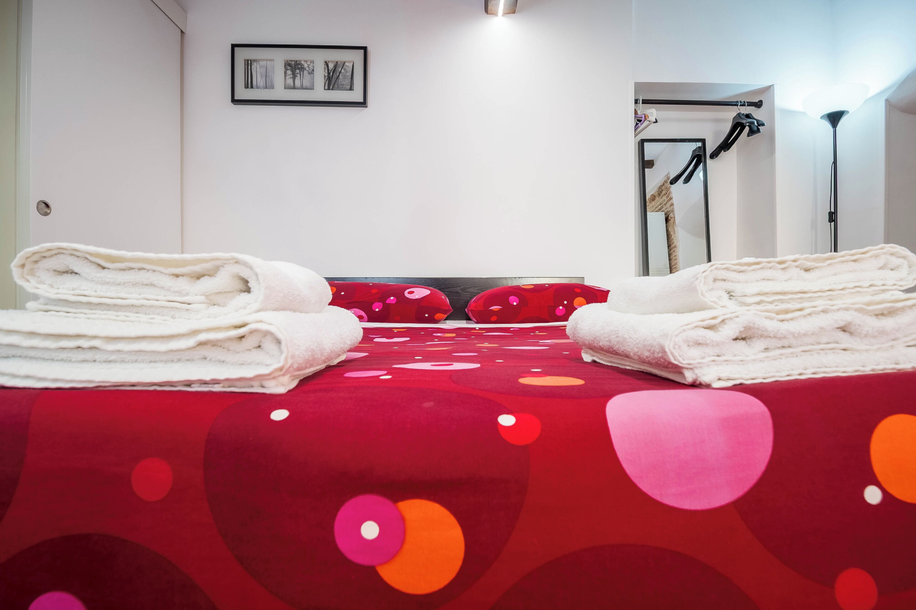 A hotel room with a red bedspread, two towels on the bed, a framed picture on the wall, and a mirror with a towel rack.