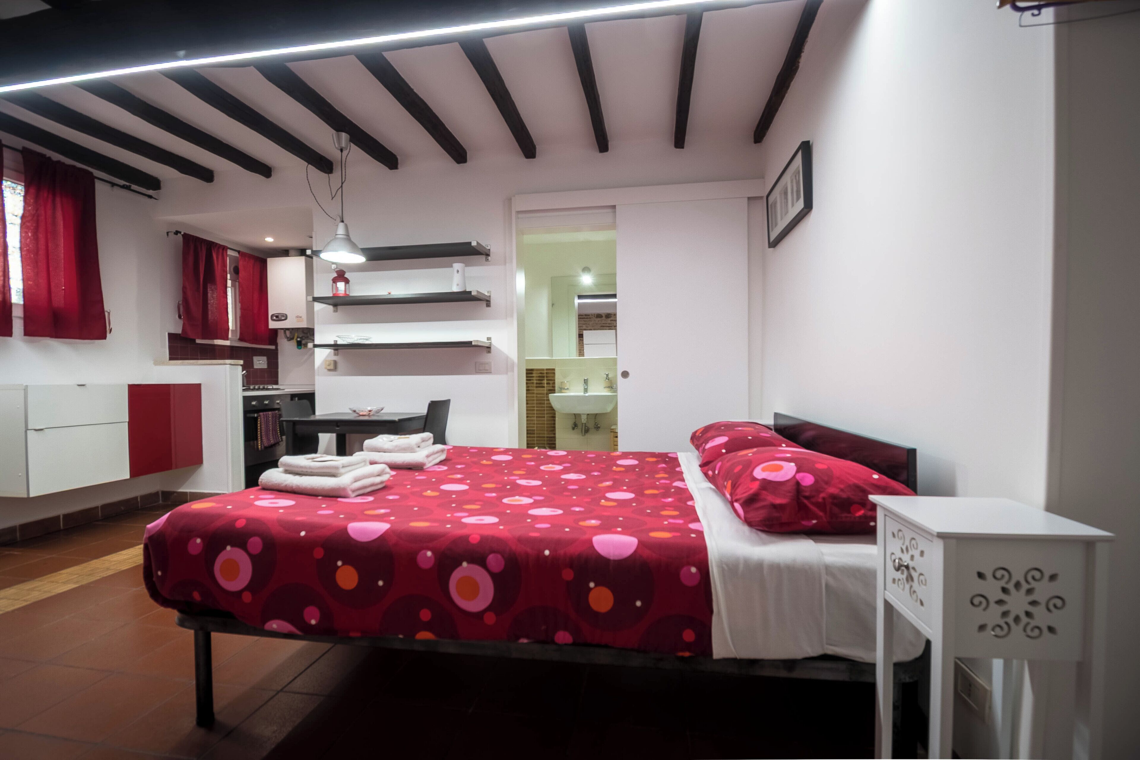A bedroom with a bed, a bedside table, a desk, a chair, a wardrobe, a hanging light, and a bathroom visible through an open door.