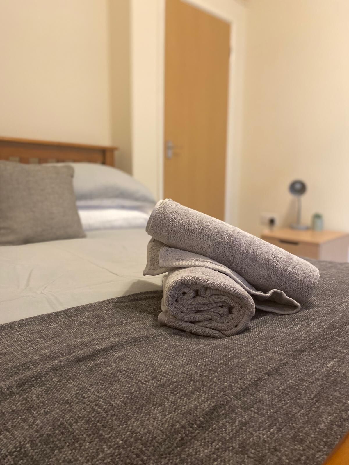 A neatly made bed with a folded towel on top, a grey blanket, and a grey pillow.