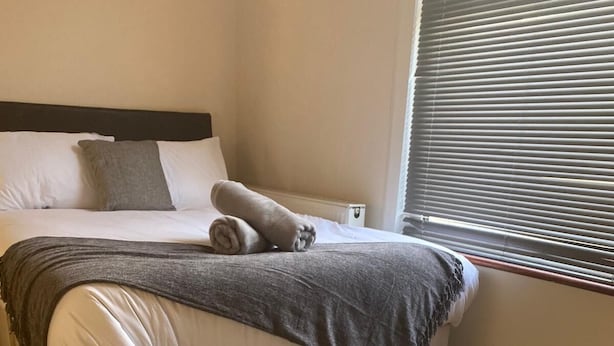 Un lit bien fait, recouvert d’une couverture grise et agrémenté d’une serviette roulée, une table de chevet blanche avec deux petites plantes grasses en pot, et une fenêtre avec des stores.