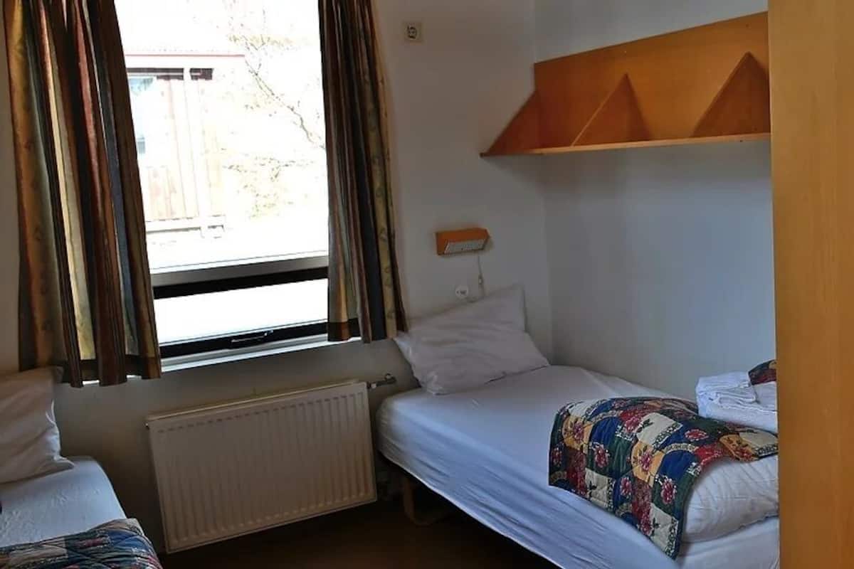 Ein kleines Zimmer mit einem Einzelbett, einem Fenster, einem Heizkörper, einem Holzschrank und einem an der Wand befestigten Regal.