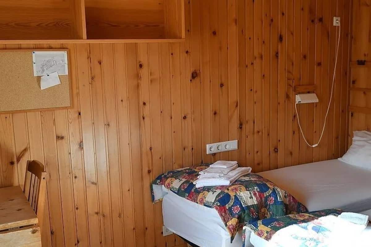 Ein Zimmer in einer Holzhütte mit zwei Betten, einem kleinen Tisch, einem Stuhl und einer Pinnwand mit Notizen.